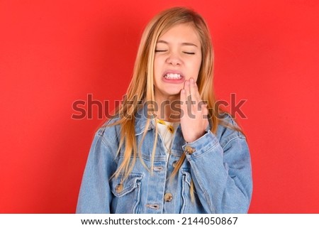 blonde little kid girl wearing denim jacket over red background  touching mouth with hand with painful expression because of toothache or dental illness on teeth.