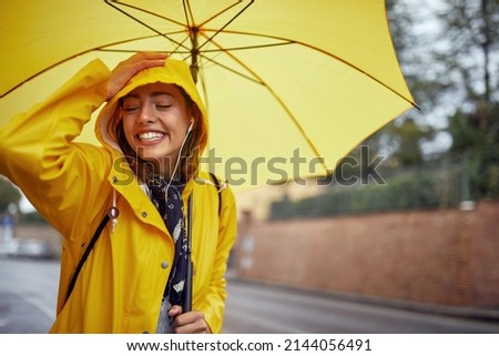 Close-up of a young cheerful woman with a yellow raincoat and umbrella who is in a good mood while walking the city on a rainy day in a relaxed manner and posing for a photo. Walk, rain, city