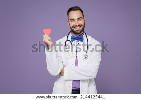 Smiling young bearded doctor man wearing white medical gown stethoscope hold red little heart isolated on violet colour wall background studio portrait. Healthcare personnel health medicine concept Royalty-Free Stock Photo #2144125441