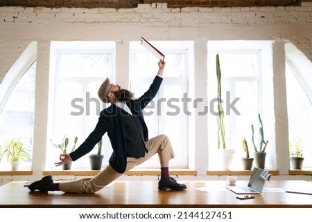 Unusual business process. Young bearded man, office clerk having fun, doing yoga on wooden table in modern office at work time with gadgets. Concept of business, healthy lifestyle, sport, hobby Royalty-Free Stock Photo #2144127451
