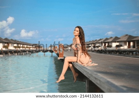 Summer vacation of Young woman relax on Maldives Resort. Romantic brunette girl in dress sitting on wooden pier by luxury water villas, enjoying ocean breeze on Paradise Island Beautiful Destination.
