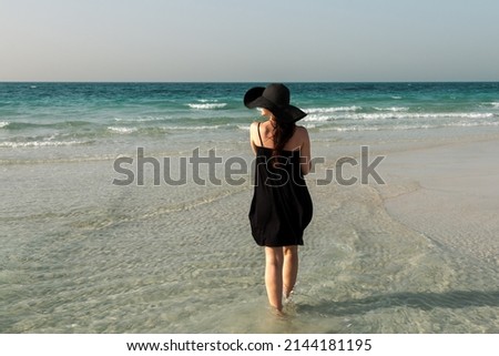 A girl in a black hat and dress stands on the background of the sea 