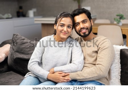Multi ethnic couple in love rent a house together. Happy Indian newlyweds moved in new apartment, sitting at the sofa in embraces, looking at the camera Royalty-Free Stock Photo #2144199623