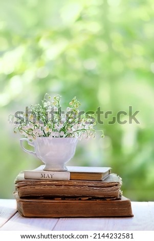 Lily of the valley flowers in cup and old books on wooden table, green blurred natural background. symbol of spring season. romantic floral composition. may month calendar