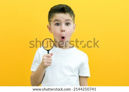 Positive curious schoolboy in white t-shirt looking at camera through magnifying glass on yellow background Royalty-Free Stock Photo #2144250741