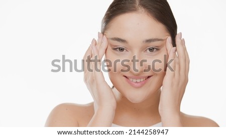 Pretty Caucasian young woman massages her temples stretching skin around eyes and smiles on white background | Droopy eyes removal concept Royalty-Free Stock Photo #2144258947