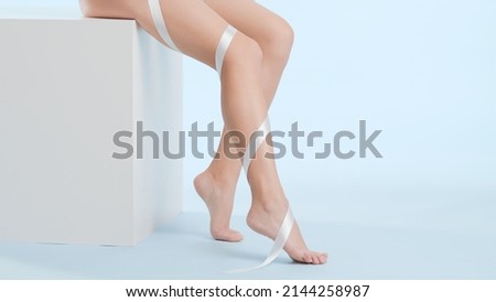Slim woman with smooth legs sits on white cube podium on pale blue background. Silk ribbon wrapped leg | Leg skin care concept Royalty-Free Stock Photo #2144258987