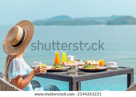 Dinner with sea view in luxury hotel. Woman in straw hat near swimming pool, eating food and enjoy ocean view. Dinner table on tropical vacation. Back view. Concept of travel, holidays, weekend. Royalty-Free Stock Photo #2144263221