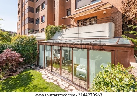 Brown aluminum and glass covered terrace in the lower part of a residential apartment building with sliding doors, decorative trees and a cobbled path