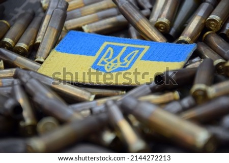 Ukrainian national flag with the coat of arms on the background of military camouflage surrounded by shells. War in Ukraine. Stop war.