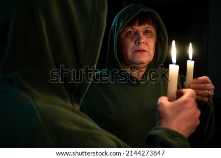 An elderly woman with a candle in her hands looks at her reflection in the mirror.