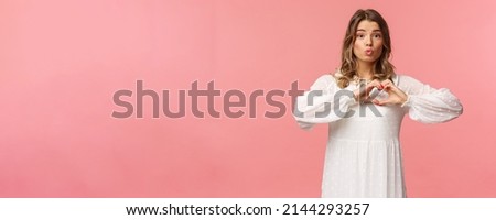 Beauty, fashion and women concept. Portrait of tender lovely young blond girl in beautiful white dress, fold lips in kiss and make heart sign near chest, standing pink background, show sympathy
