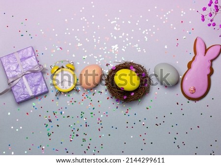 Easter composition - a nest with painted yellow and blue eggs, retro alarm clock, lilac gift box, gingerbread cookies in the shape of a purple Easter bunny top view. Time to celebrate Easter concept.