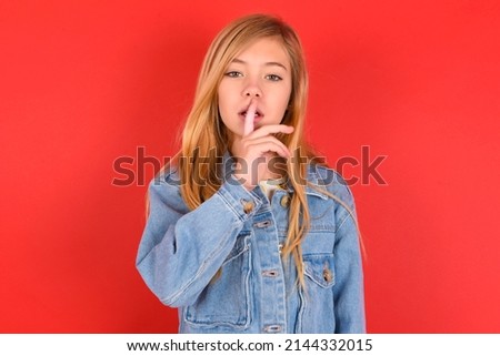 blonde little kid girl wearing denim jacket over red background   makes silence gesture, keeps finger over lips. Silence and secret concept.