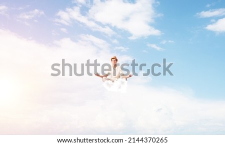 Man in white clothing keeping eyes closed and looking concentrated while meditating on clouds in the air with cloudy skyscape on background.