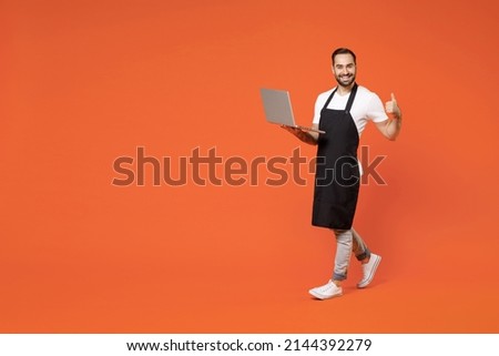 Full length young man barista bartender barman employee in black apron white t-shirt work in coffee shop use laptop pc computer thumb up gesture isolated on orange background. Small business startup