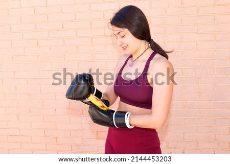 A young Caucasian woman is wearing black boxing gloves and trying to peel a banana. In the background is a brick wall.