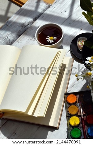 Open notebook, cup of tea and watercolor paints on white wooden background