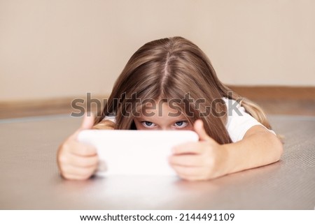 Little girl child uses smartphone, looks at screen, plays or watches cartoon and lies on floor. Kid with Gadget, mockup Royalty-Free Stock Photo #2144491109