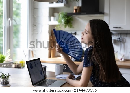 Woman cools herself with waver, lean at table with laptop distracted from work feels exhausted by intense heat in summer hot day inside kitchen without climate control, need air conditioner concept Royalty-Free Stock Photo #2144499395