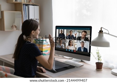 Young woman sit at desk in front of pc, looks at screen, wave hand greeting diverse friends, multinational colleagues start or finish group video call. Virtual meeting, remote communication concept Royalty-Free Stock Photo #2144499407