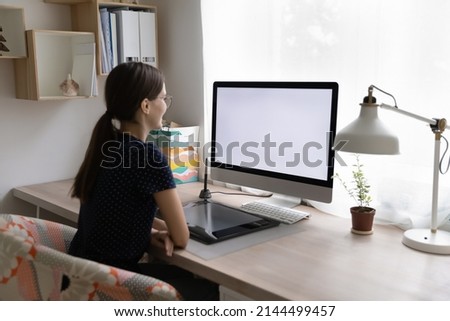 Rear view young woman sit at workplace desk with digital graphic tablet, staring at computer white mock up screen looks interested working or learns professional photo or video editor program concept
