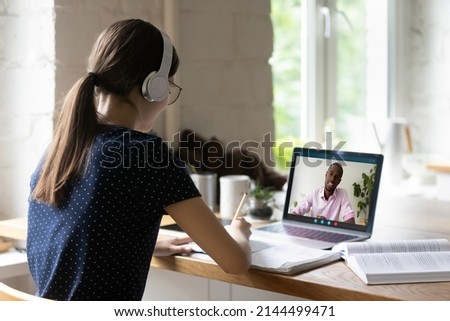 Rear back view student girl sit at desk with laptop, wear headphones learns english remotely make call take part in class with African tutor. E-learn, tuition, gain knowledge use modern tech concept Royalty-Free Stock Photo #2144499471