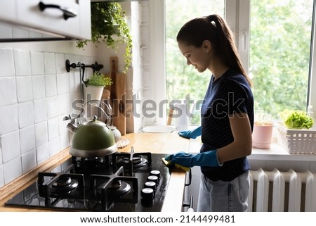 Young housewife wear rubber protective gloves standing in cozy kitchen cleans worktop table use rag and effective detergent products. Household, cleaning services, domestic work, housekeeping concept Royalty-Free Stock Photo #2144499481