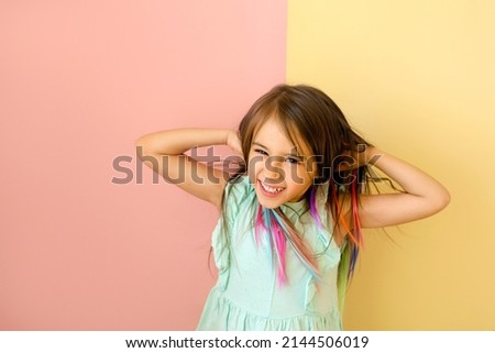 Cheerful cute funny girl with bright multi colored strands of hair laughs merrily and straightening hair with hands. Child with artificial locks of hair on a yellow-pink studio background. 