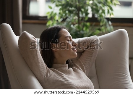 Relaxed calm positive millennial girl resting in comfortable armchair, leaning back with sleepy closed eyes, enjoying relaxation, home leisure, breathing fresh air for stress relief, dreaming Royalty-Free Stock Photo #2144529569
