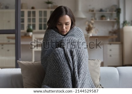 Stressed sad wrapped in plaid young woman feeling cold, ill, sick, suffering from fever, virus, trying to warm, sitting on couch at home. Frustrated shocked girl going through depression Royalty-Free Stock Photo #2144529587