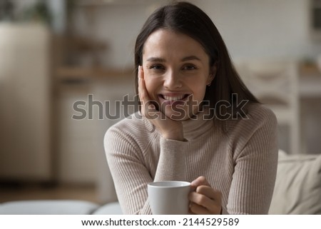 Happy pretty girl drinking morning tea, cocoa, chocolate, holding cup of hot beverage, enjoying home leisure, looking at camera, smiling, relaxing. Head shot portrait