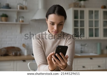 Focused smartphone user girl chatting on social media in home kitchen, holding mobile phone, typing text message, browsing internes. Young woman using online app, service on cellphone