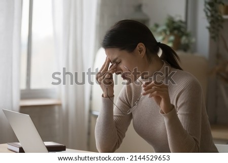 Tired upset laptop user girl overworking at home, sitting at table with computer at home workplace, holding glasses, touching closed eyes, suffering from weak eyesight, vision problems Royalty-Free Stock Photo #2144529653