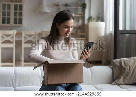 Happy young customer woman receiving purchase from internet store, using app on smartphone, giving feedback to shop, unpacking parcel from online shop, using cellphone, making delivery order Royalty-Free Stock Photo #2144529655