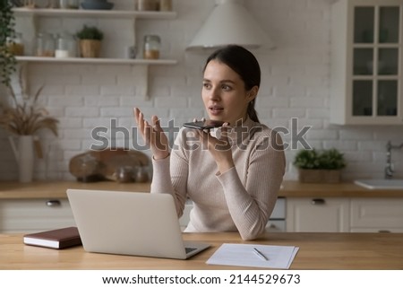Busy serious freelance professional woman talking on speaker on smartphone at home workplace. Business woman, entrepreneur, remote employee giving voice command to virtual assistant on cell