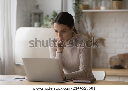 Thoughtful serious freelance worker woman working from home kitchen, using laptop, thinking over email letter, online project. Focused student girl studying on internet sitting at computer on table Royalty-Free Stock Photo #2144529681