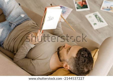 A young artist holds a sketchbook in his hands. A man in a khaki long sleeve T-shirt is resting on the sofa in his workshop. Rest, search for inspiration, artist in the studio.