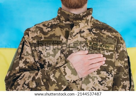 Ukrainian patriot soldier in military uniform holds a hand on a heart against the background of a yellow and blue flag