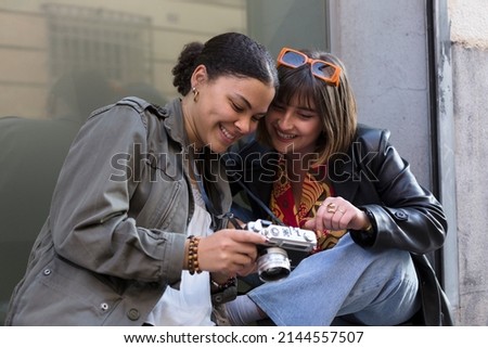 Two smiling multiethnic friends looking at pictures on camera. Concept of having fun time outdoors.