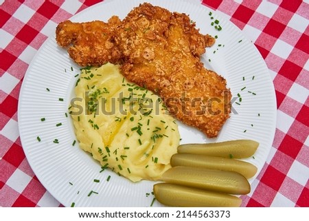Close up picture of deep fried chicken schnitzel with mashed potatoes and pickled sour cucumbers sprinkled with minced chives served on the white rustic plate. Traditional dish from Czech Republic. 