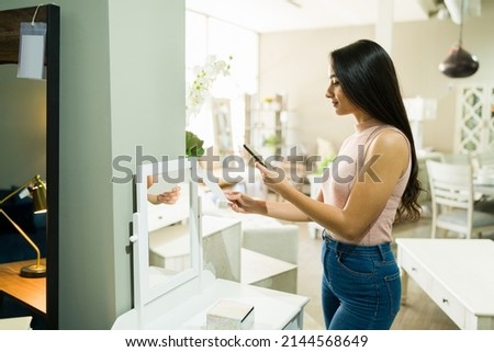 Beautiful young woman looking at the price of a vanity mirror and taking a picture with a smartphone at the furniture store