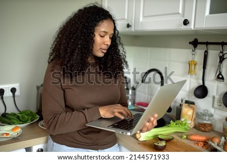 Horizontal picture of dark-skinned female in brown clothes standing against kitchen counter full of vegetables and food, holding laptop, searching for recipe, cooking breakfast on Sunday morning