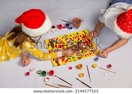 Boy and girl in red santa claus hats lie on the floor and draw a Christmas picture at home. Children paint a gingerbread man with paints.