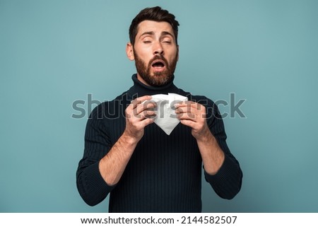 Influenza season. Unhealthy flu-sick man sneezing loudly in tissue, feeling unwell with runny nose, caught cold or allergy symptom. studio shot isolated on blue background  Royalty-Free Stock Photo #2144582507