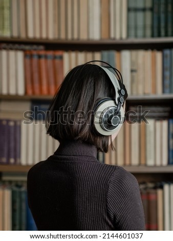 Woman in headphones listening to audiobooks on background of library shelves with paper books.