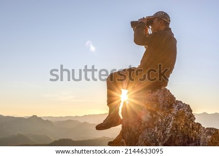 Male hikers holding binoculars sitting on top of the rock mountain at sunset background.