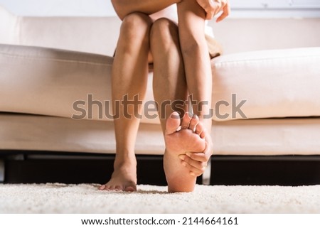 Foot pain, Asian woman sitting on sofa feeling pain in her foot at home, female suffering from feet ache use hand massage relax muscle from soles in home interior, Healthcare and podiatry medical