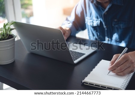 Close up of man working on laptop computer and hand with a pen writing on notebook on office table, online study, e-learning, digital education, telecommuting, work remotely concept