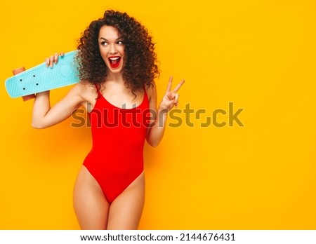 Young beautiful smiling woman posing near yellow wall in studio.Sexy model in red swimwear bathing suit.Positive female with afro curls hairstyle. Holding penny skateboard. Happy and cheerful Royalty-Free Stock Photo #2144676431